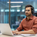 Young professional wearing headphones working on laptop in modern office environment while learning how to become a legal transcriptionist
